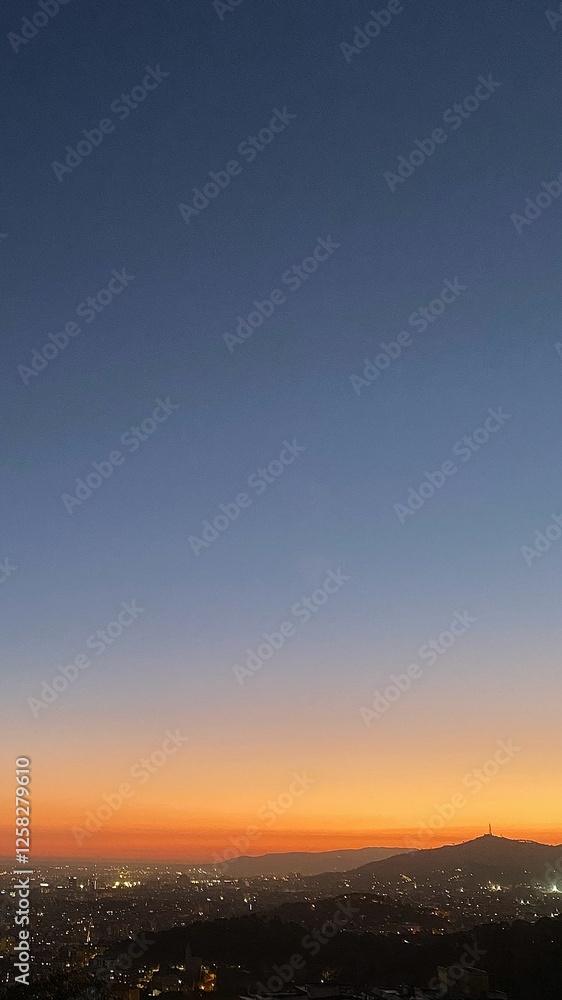 Fototapeta premium Cityscape at dusk with a gradient sky transitioning from deep blue at the top to warm orange near the horizon. City lights glow as the sun sets, with mountains silhouetted in the distance