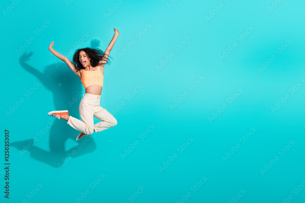 Fototapeta premium Excited young woman jumping joyfully on a vibrant turquoise background, expressing energy and happiness in casual summer fashion.