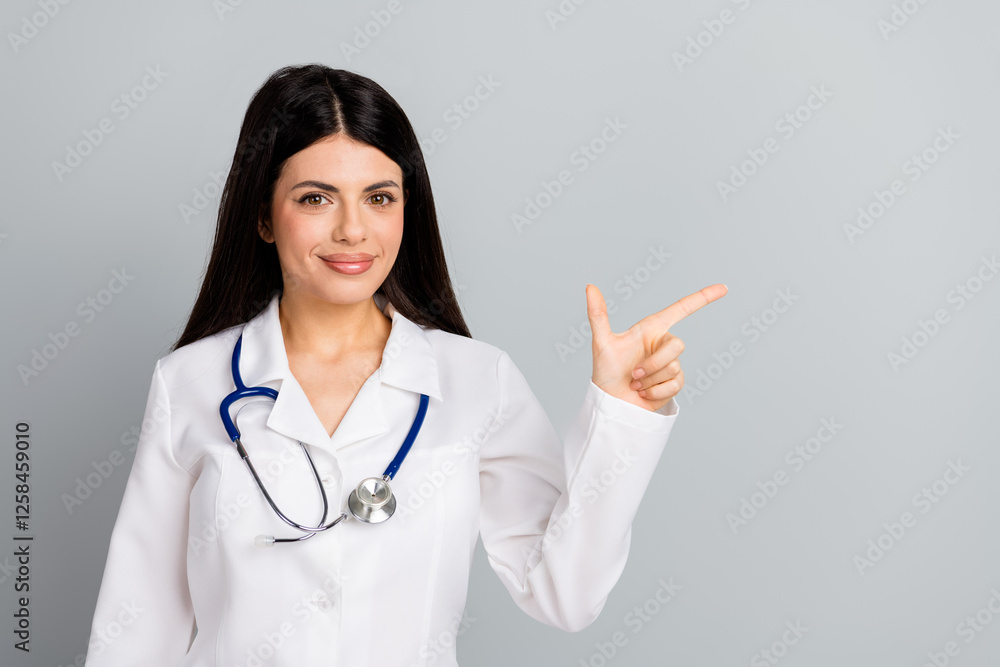 Confident female doctor wearing white coat and stethoscope pointing at something with a smile against a gray background Confident female doctor wearing white coat and stethoscope pointing at something with a smile against a gray background