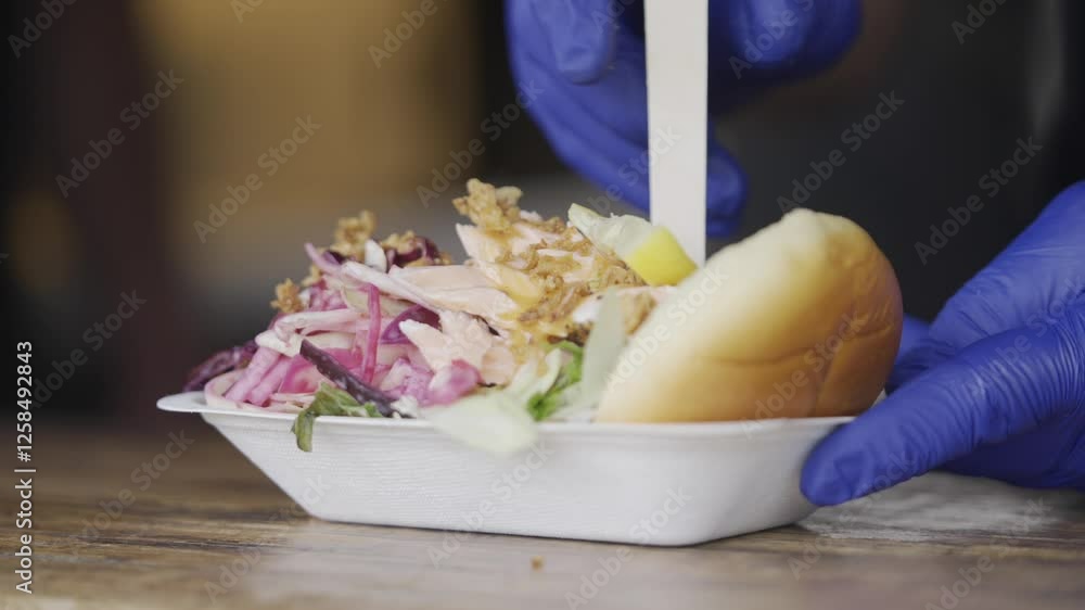 A person is holding a sandwich and salad in a white container. The person is wearing blue gloves A person is holding a sandwich and salad in a white container. The person is wearing blue gloves