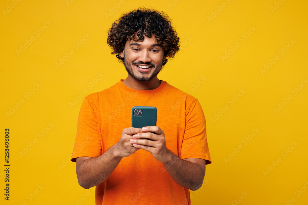 Young smiling happy student Indian man he wear orange t-shirt casual clothes hold in hand use mobile cell phone chatting online isolated on plain yellow background studio portrait. Lifestyle concept.