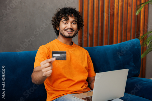 Young Indian man in orange casual clothes sits on blue sofa couch use laptop pc computer hold credit bank card shopping online stay at home hotel flat rest relax spend free time in living room indoor. photo