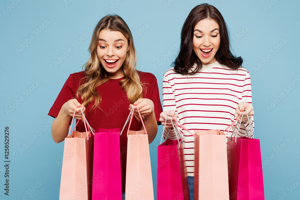 Young fun couple two friends women wearing red casual clothes together hold in hand open paper package bags after shopping isolated on plain blue background studio. Black Friday sale buy day concept. Young fun couple two friends women wearing red casual clothes together hold in hand open paper package bags after shopping isolated on plain blue background studio. Black Friday sale buy day concept.