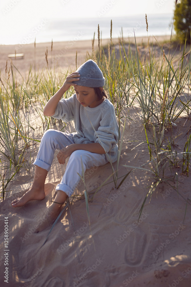 A fashionable seven-year-old boy sit along a sandy beach by the sea in autumn in a sweater and hat at sunset.