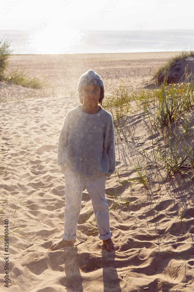 A fashionable seven-year-old boy stay along a sandy beach by the sea in autumn in a sweater and hat at sunset.