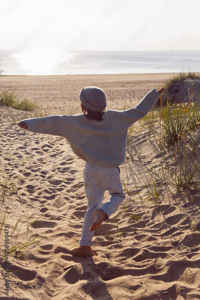 A fashionable seven-year-old boy runs along a sandy beach by the sea in autumn in a sweater and hat at sunset.