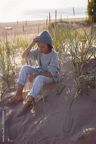 Obraz na płótnie A fashionable seven-year-old boy sit along a sandy beach by the sea in autumn in a sweater and hat at sunset. w salonie