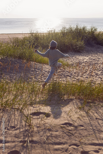 Obraz na płótnie A fashionable seven-year-old boy runs along a sandy beach by the sea in autumn in a sweater and hat at sunset. w salonie