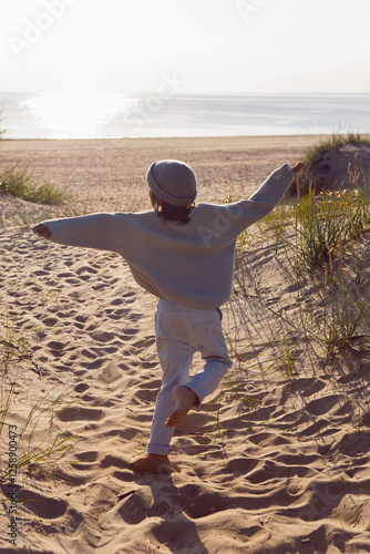 Obraz na płótnie A fashionable seven-year-old boy runs along a sandy beach by the sea in autumn in a sweater and hat at sunset. w salonie