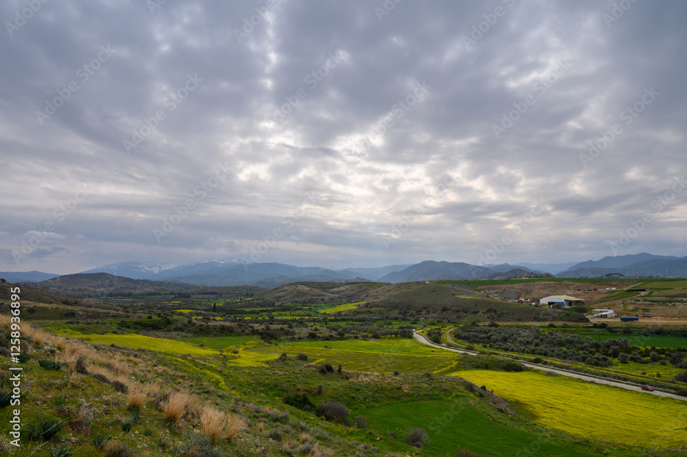 Lush countryside stretches under a cloudy sky with vibrant fields and distant mountains in the background Lush countryside stretches under a cloudy sky with vibrant fields and distant mountains in the background
