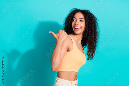 Portrait of a cheerful young woman making a thumb gesture against a teal background, showcasing a stylish casual outfit photo