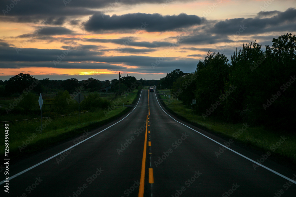 Fototapeta premium Setting sun on a two-lane blacktop highway in the country with a dramatic sky