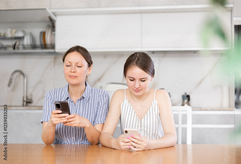 Happy mother with adult daughter writing messages on smartphone screen while sitting at table in kitchen Happy mother with adult daughter writing messages on smartphone screen while sitting at table in kitchen