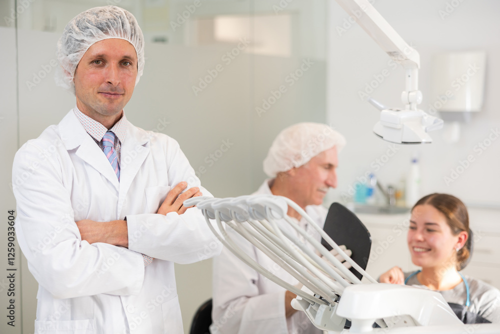 In dentists office, man dentist before start of appointment working day. Positive medical specialist welcome clinics clients. Doctor enjoy working in profession. Patient treatment in background In dentists office, man dentist before start of appointment working day. Positive medical specialist welcome clinics clients. Doctor enjoy working in profession. Patient treatment in background