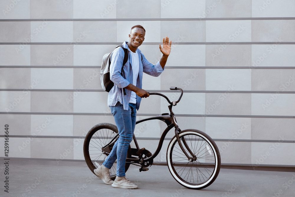 Positive millennial man with modern bike waving near brick wall outdoors Positive millennial man with modern bike waving near brick wall outdoors