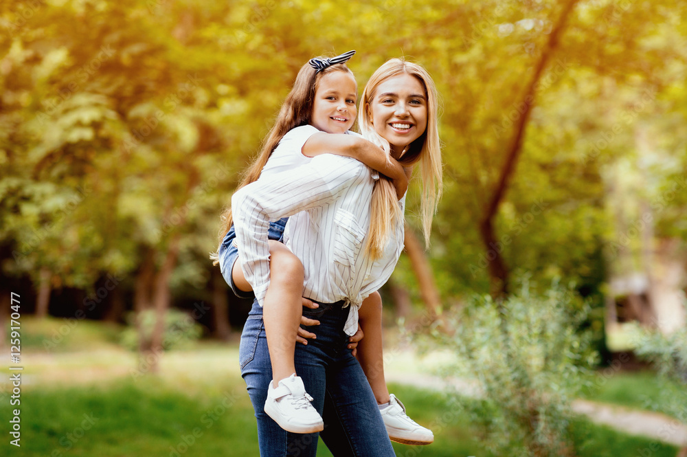 Fototapeta premium Joyful Mom Carrying Little Daughter Piggyback Having Fun Playing In Park Outdoors. Happy Motherhood Concept