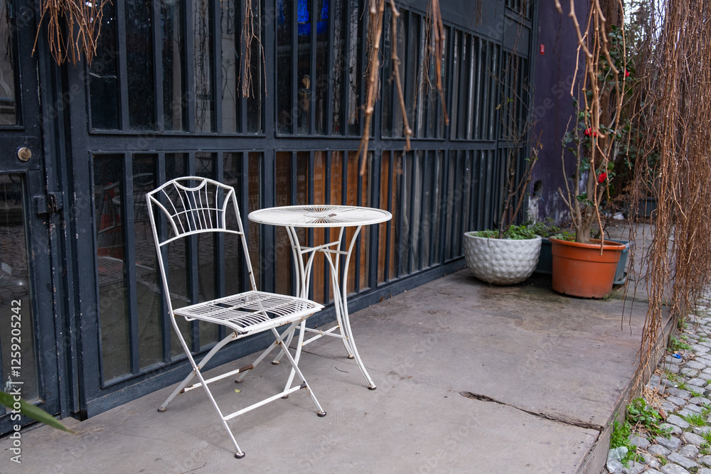 Solitary white metal table and chair set against a minimalist industrial building. Outdoor seating area with a white metal table and chair, situated in front of a grimy industrial building Solitary white metal table and chair set against a minimalist industrial building. Outdoor seating area with a white metal table and chair, situated in front of a grimy industrial building