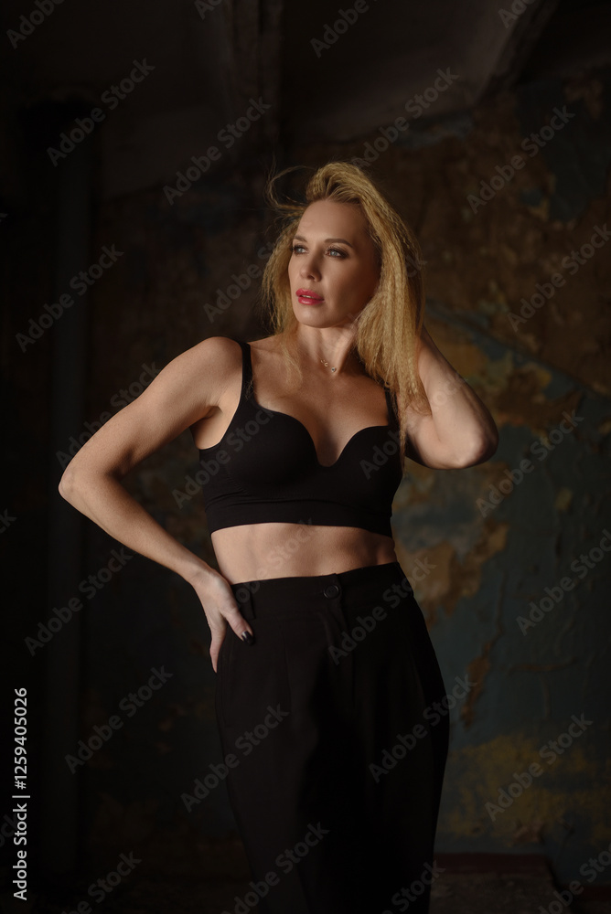 Fototapeta premium Beautiful blonde woman posing by the window in black top and trousers in old corridor.