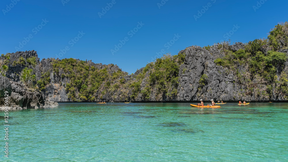 Obraz premium Canoes with tourists sail along the turquoise lagoon. Green vegetation on the steep slopes of coastal karst cliffs. Clear blue sky. Philippines. Palawan. Big Lagoon. Bacuit bay