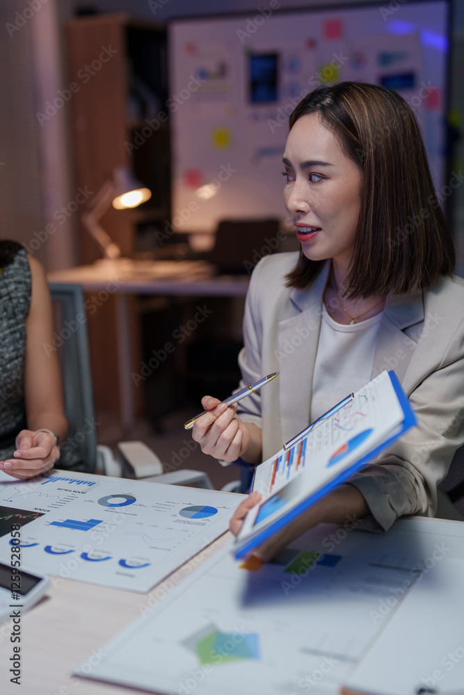 Fototapeta premium Asian businesswomen engaging in a meeting, discussing marketing strategies while analyzing financial statistics and pointing at charts in a corporate office during the night