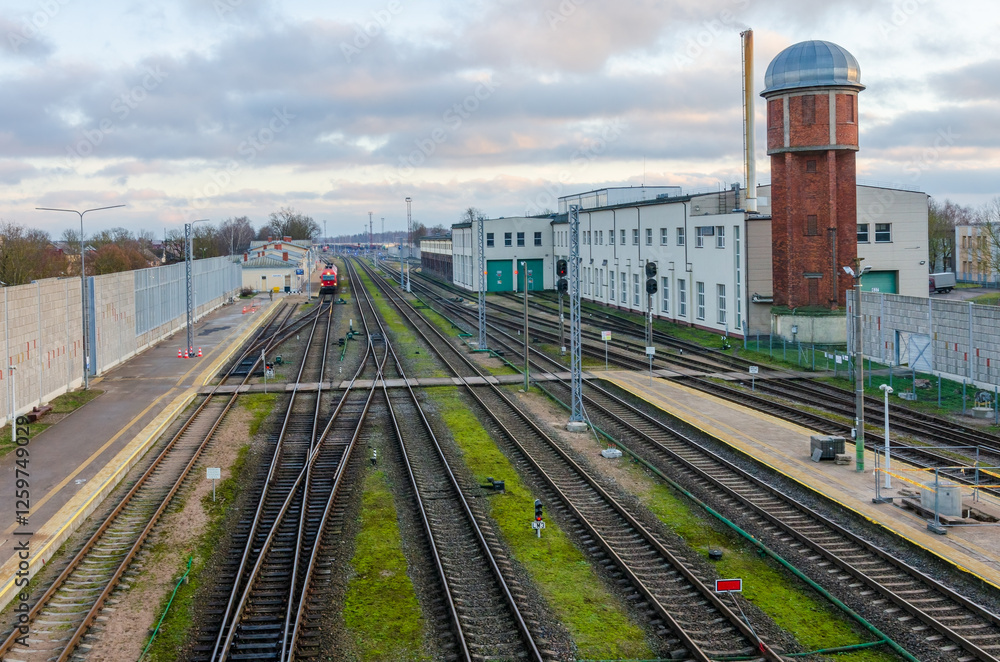 Fototapeta premium Railway station with multiple tracks, train, and historic water tower at dusk. Train station scene, calm atmosphere, wide-angle shot, elevated perspective, industrial setting, multiple railway tracks,