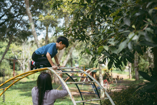 Foto A young 8 years old boy climbing a jungle gym with the assistance of his mother in a park, surrounded by green trees and natural light