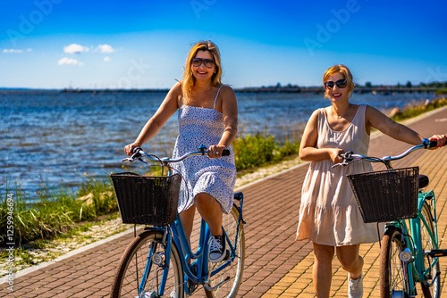 Two mature women: one riding bicycle, second walking with bike on bicycle path by sea on beautiful sunny day in summertime. Front view photo