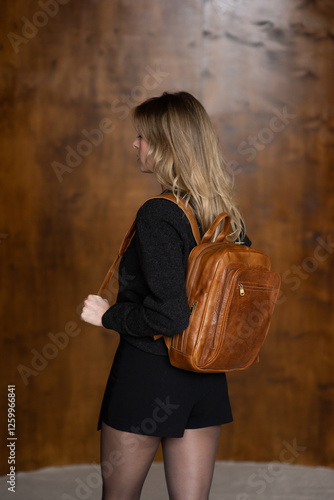 A girl in a warm sweater, a black short skirt and nylon tights is posing with a brown backpack.