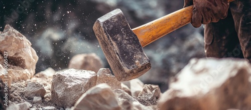 Close-up of a weathered construction worker's hand gripping a heavy sledgehammer poised above large rugged rocks in a gritty quarry setting