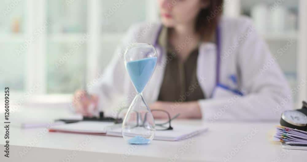 Woman doctor in uniform makes records sitting at workplace with hourglass. Female medic with stethoscope creates patient history in hospital Woman doctor in uniform makes records sitting at workplace with hourglass. Female medic with stethoscope creates patient history in hospital