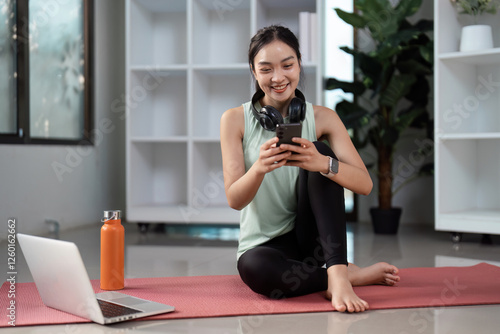 Cheerful Asian woman using smartphone while sitting on yoga mat at home, promoting wellness and a balanced lifestyle. photo