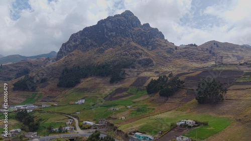 Aerial view revealing rock formation towering over patchwork farmlands and small rural settlement within mountainous landscape