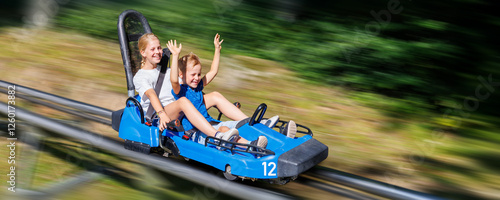 Pair of cute laughing kids enjoy have fun riding alpine roller coaster cart together at Harz nature park on sunny holidays summer day. Children summer outdoors travel active sports adventure