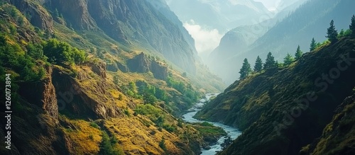 Lush green river canyon nestled between majestic mountains with selective focus on glistening water and distant fog-covered peaks in morning light