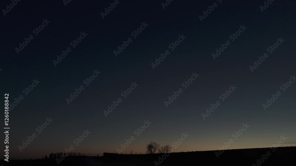 serene countryside road at dawn. The dark silhouettes of leafless trees stand against a deep blue sky, which gradually transitions to a warm orange glow near the horizon. The setting appears peaceful serene countryside road at dawn. The dark silhouettes of leafless trees stand against a deep blue sky, which gradually transitions to a warm orange glow near the horizon. The setting appears peaceful
