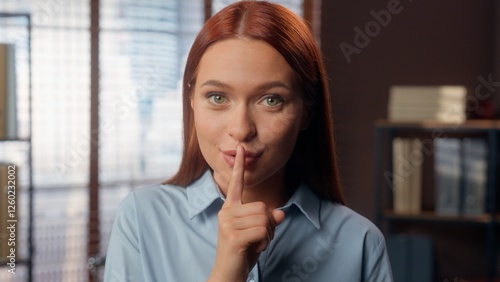 Smiling Caucasian business woman girl female businesswoman employer manager in office hold index finger near lips mouth hush silence quiet gesture secret secrecy information silent looking at camera