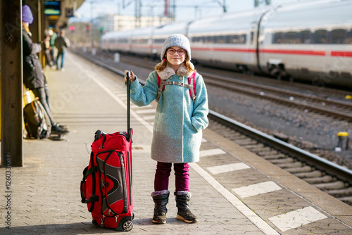 Little happy girl with glasses and a winter outfit standing on a train platform, holding a red rolling suitcase while waiting for a high-speed train photo