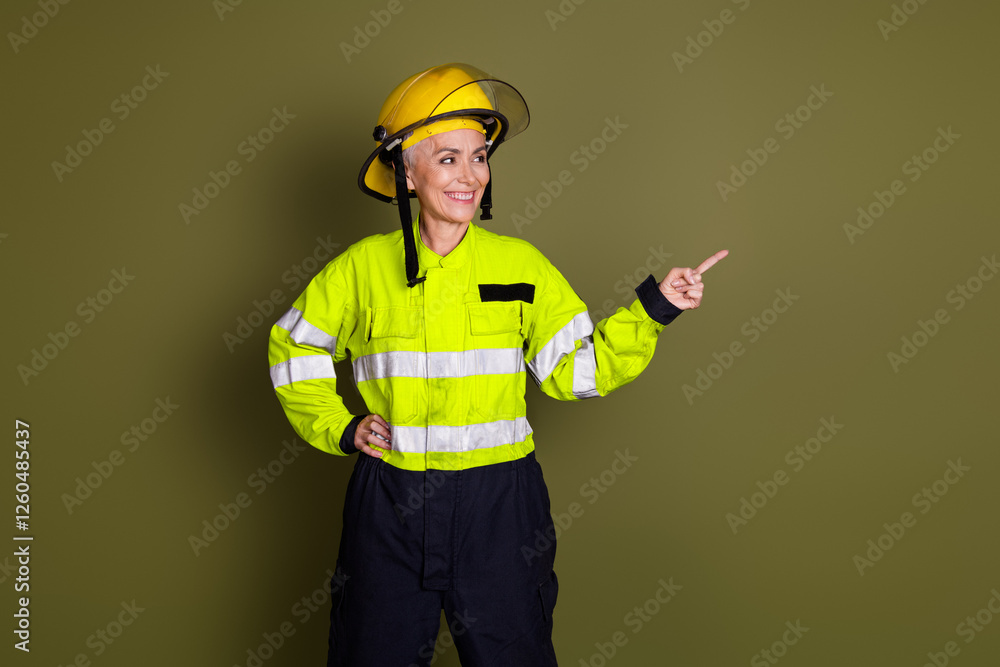 Confident woman in safety gear pointing while smiling, wearing a protective helmet and high-visibility uniform against a khaki background Confident woman in safety gear pointing while smiling, wearing a protective helmet and high-visibility uniform against a khaki background