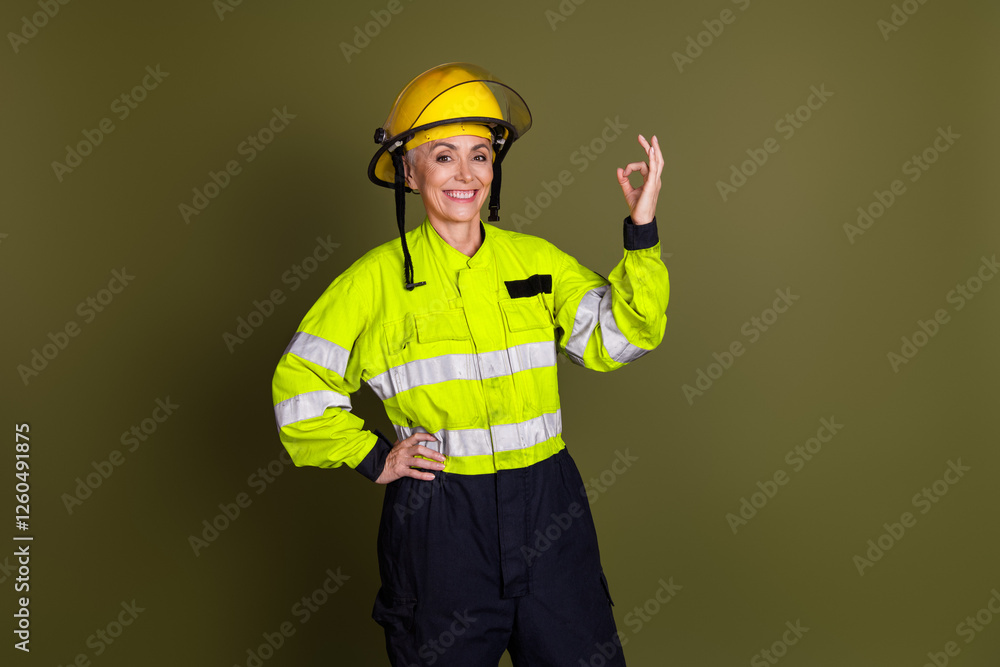 Confident mature female worker in high visibility workwear, protective helmet showing okay gesture on khaki background Confident mature female worker in high visibility workwear, protective helmet showing okay gesture on khaki background