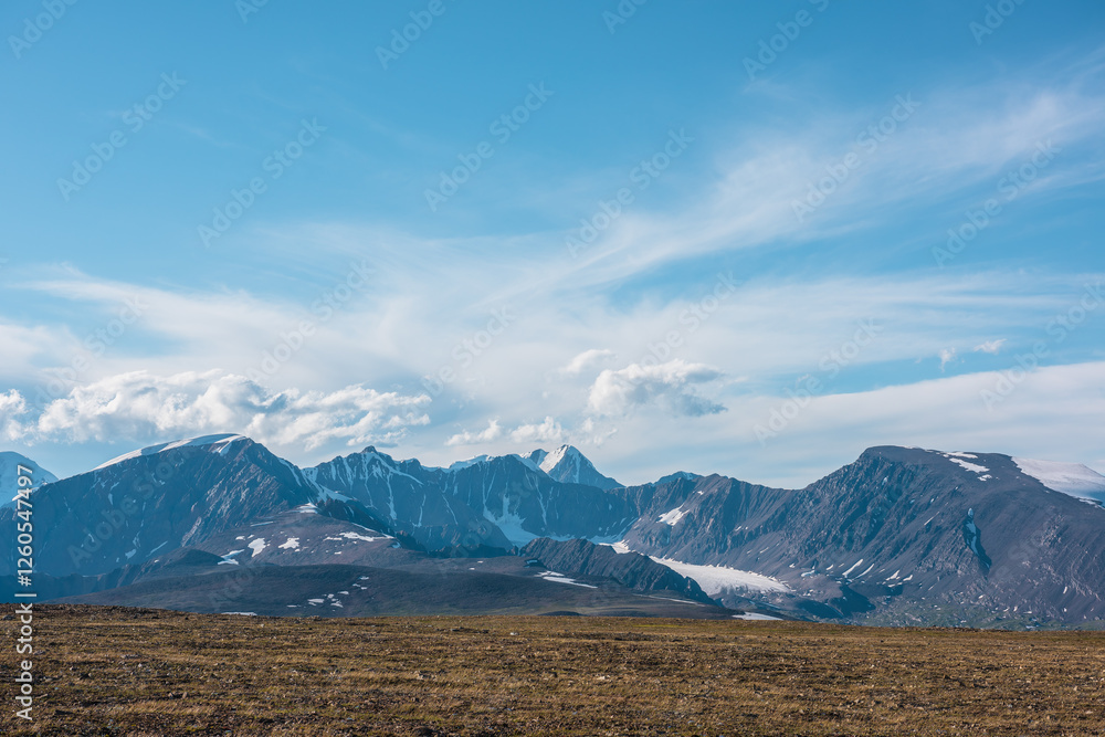 Top view from sunlit grassy stony plain of pass to large rocky mountain range with glacier and high snow pointy peak in bright sun under clouds in blue sky. Big snowy peaked top far away in sunny day. Top view from sunlit grassy stony plain of pass to large rocky mountain range with glacier and high snow pointy peak in bright sun under clouds in blue sky. Big snowy peaked top far away in sunny day.