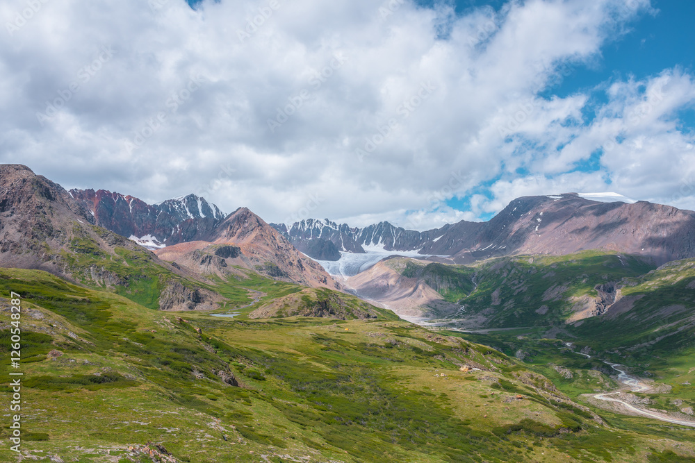 Colorful vast scenery of wide green alpine valley with panoramic view to large snow-capped top, sharp rockies, rocky pointy peak, snowy mountain range and big glacier tongue far away under cloudy sky. Colorful vast scenery of wide green alpine valley with panoramic view to large snow-capped top, sharp rockies, rocky pointy peak, snowy mountain range and big glacier tongue far away under cloudy sky.