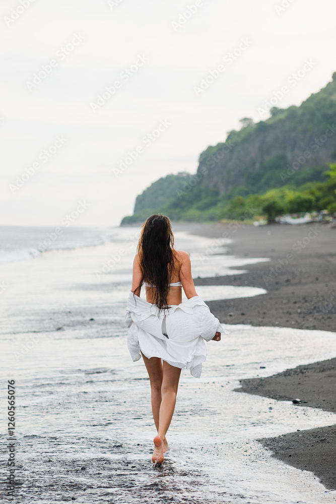 Barefoot Woman in White Loose Shirt Walking on Black Sand Beach, Bali, Indonesia. Barefoot Woman in White Loose Shirt Walking on Black Sand Beach, Bali, Indonesia.