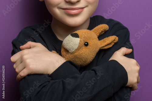 Adorable child with arms crossed, hugging a cute kangaroo plushie on a purple background. Represents themes of love, care, and the warmth of childhood memories. photo