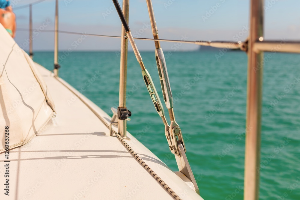 Fototapeta premium A close-up of a sailboat deck with metal rigging, ropes, and turquoise ocean water in the background. The warm sunlight highlights the details, evoking themes of sailing, adventure, and ocean travel