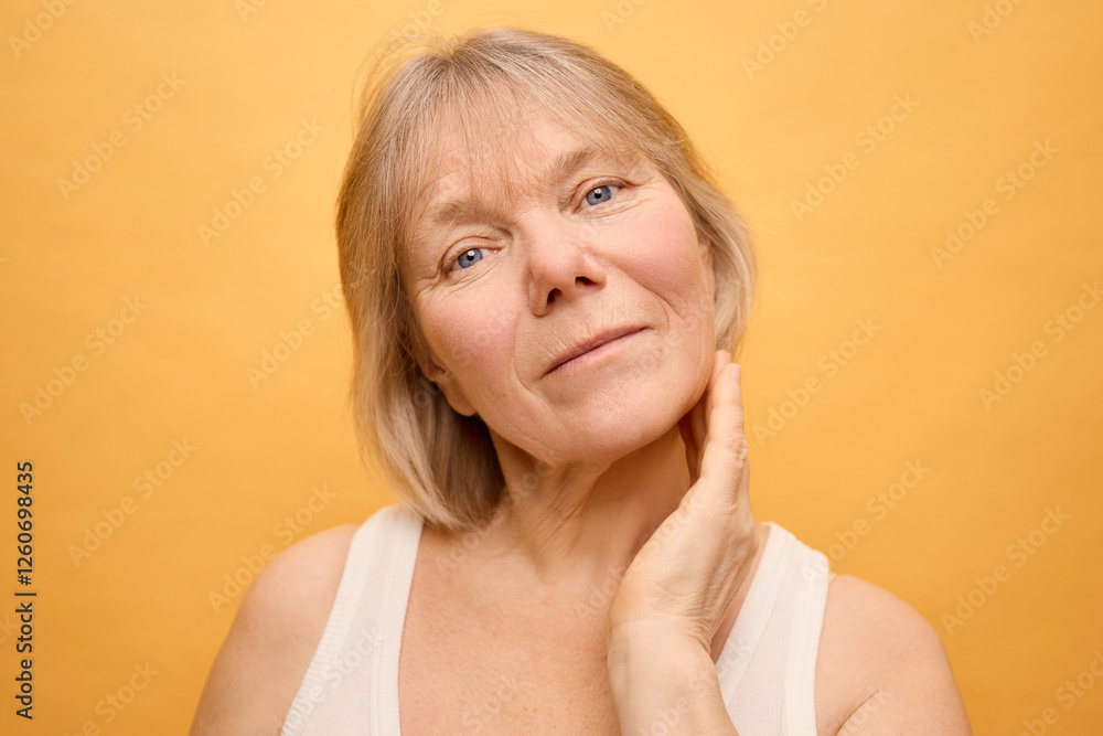 A mature woman with light blonde hair and bright blue eyes poses gracefully, lightly touching her neck. She presents a warm smile, highlighting her natural beauty with a cheerful yellow backdrop A mature woman with light blonde hair and bright blue eyes poses gracefully, lightly touching her neck. She presents a warm smile, highlighting her natural beauty with a cheerful yellow backdrop