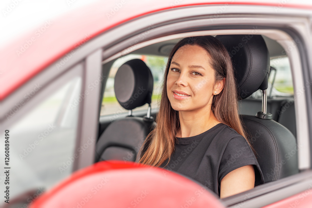 A young woman with long brown hair and a bright smile sits in the drivers seat of a red car, looking outside the open window with a cheerful and confident expression.