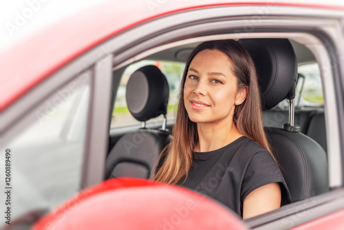 Obraz na płótnie A young woman with long brown hair and a bright smile sits in the drivers seat of a red car, looking outside the open window with a cheerful and confident expression. w salonie