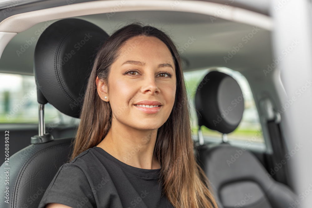 A young beautiful woman with long brown hair and a warm smile sits in the driver's seat of a modern car, looking directly at the camera with confidence.