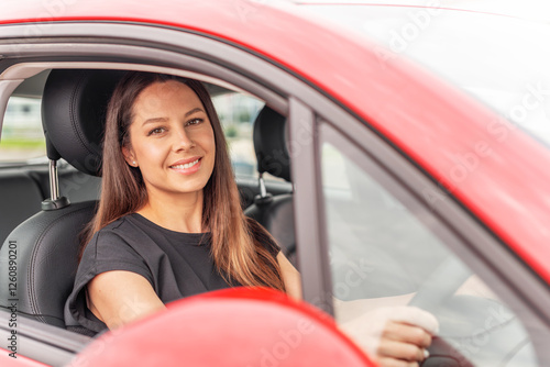 Obraz na płótnie A young beautiful woman with a bright smile and long brown hair sits confidently in the drivers seat of a red car, holding the steering wheel and looking outside with a happy expression. w salonie