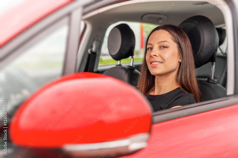 A young beautiful woman with long brown hair and a confident smile sits in the drivers seat of a red car, looking through the open window while holding the steering wheel.